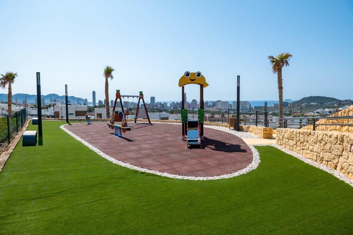 Ein Spielplatz mit Schaukeln und Rutsche, umgeben von einer Steinmauer und Palmen, mit einer Stadtlandschaft im Hintergrund.