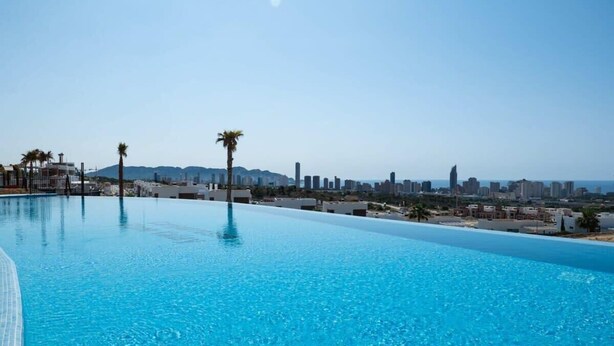 A clear blue infinity pool with a city skyline in the background. - Casa la Vista • 2 Bedroom • Sunny terrace • Camporrosso Stone Valley Resort (Finestrat)
