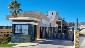 A modern building with a gated entrance, a small office-like structure labeled 'Calle Brasil 2', and a palm tree in the background. - Casa la Vista • 2 Bedroom • Sunny terrace • Camporrosso Stone Valley Resort (Finestrat)