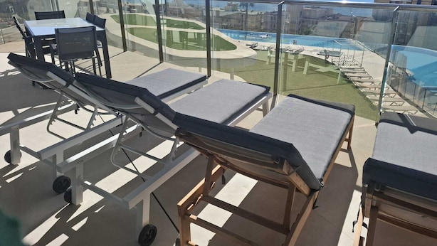 Een balkon met ligbedden, een zwembad en uitzicht op zee en de stad.