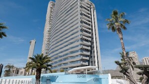 Een hoog, modern hotel met een gebogen gevel, diverse balkons en een zwembadgedeelte met de naam 'SUNSET'.