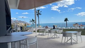 Een terras met tafels en stoelen, met uitzicht op een strand en de zee.