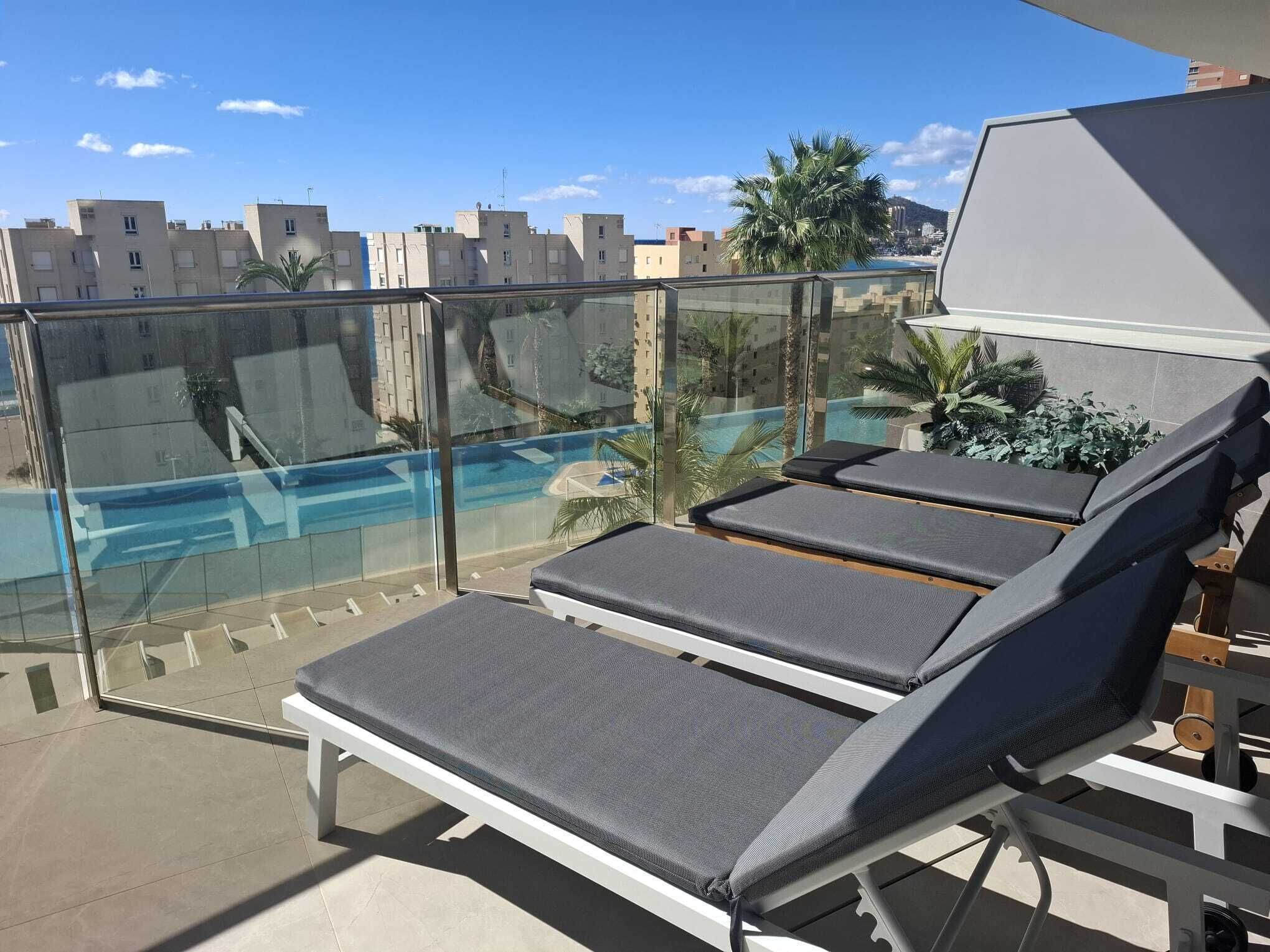 Un balcon avec deux fauteuils de relaxation, une balustrade en verre et une vue sur une piscine et des immeubles.