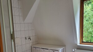 A bathroom with a white toilet, a shower with a glass door, a white vanity with storage baskets, and a window with a view of greenery. - Vacation apartment Bergblick (Königstein)