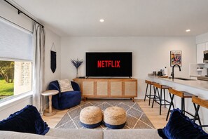 A modern living room with a sofa, ottomans, a TV displaying 'NETFLIX', a kitchen area, and a view of the outdoors. - Zilker Bungalow, Pet Friendly (Austin)