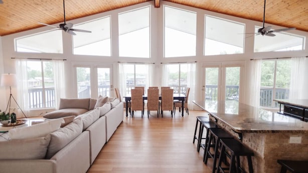 A spacious living area with a high ceiling, large windows, a sofa, dining table, chairs, and a kitchen counter.