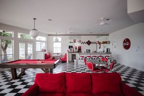 A room with a pool table, red seating, a dining area with a checkered floor, and a kitchen area with a Coca-Cola sign.