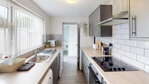 A modern kitchen with a built-in oven, microwave, and sink. There is a window with blinds, a plant, and a utensil holder on the counter. - City Centre-university Home From Home (Sunderland)