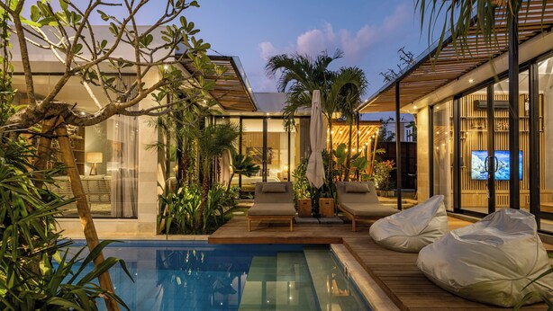 Un área de piscina moderna al aire libre con sillones de descanso y pufs, rodeada de plantas tropicales y una terraza de madera.