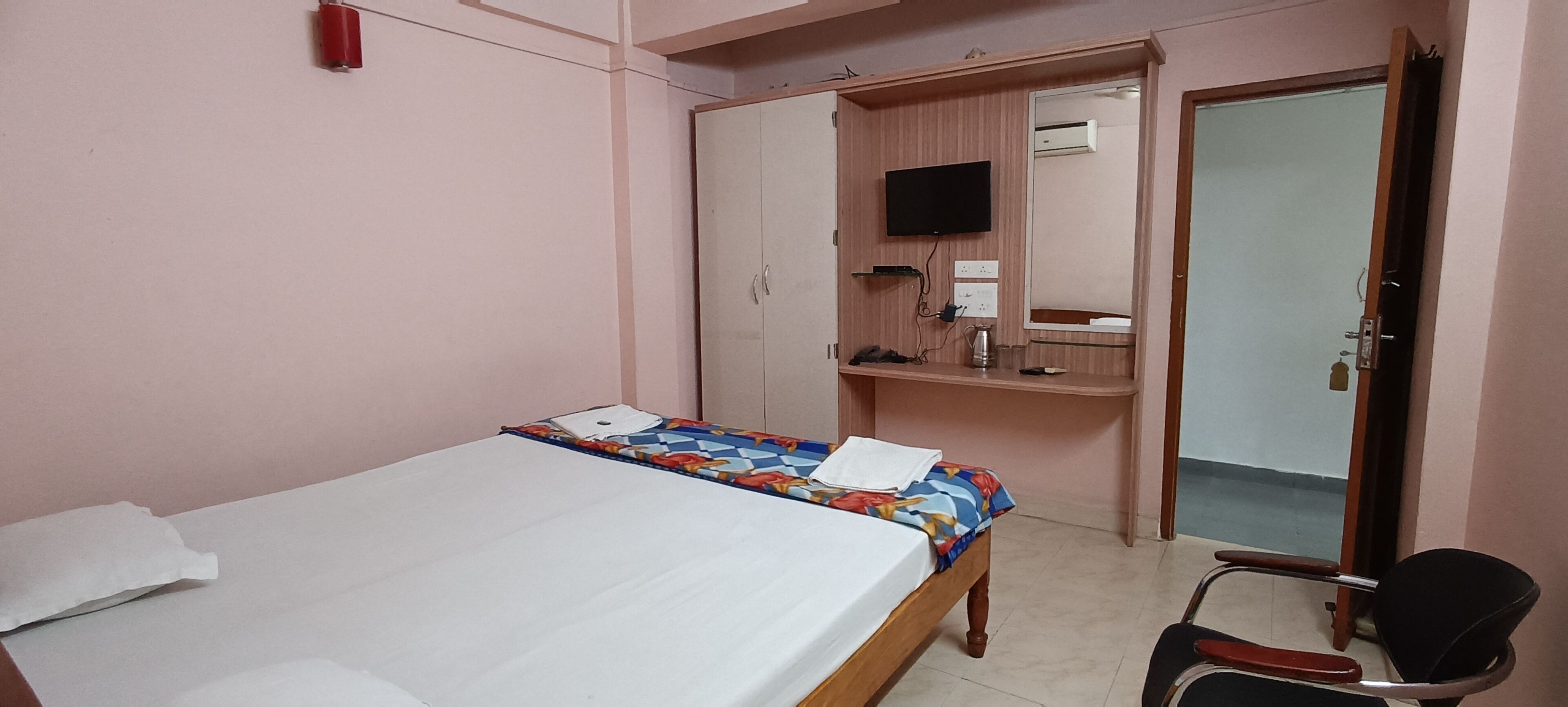 A bedroom with a bed, a desk, a chair, a wardrobe, a television, and an air conditioning unit.
