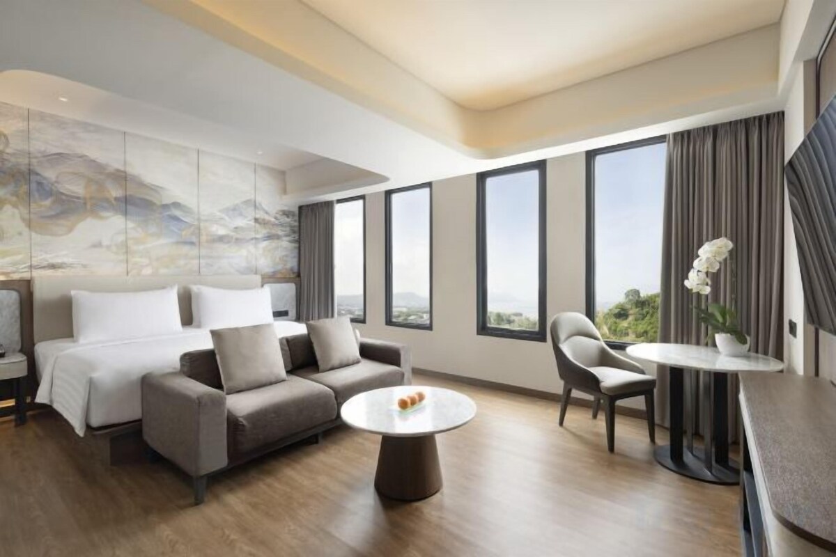 Quarto de hotel moderno com cama, sofá, mesa de jantar e grandes janelas com vista.