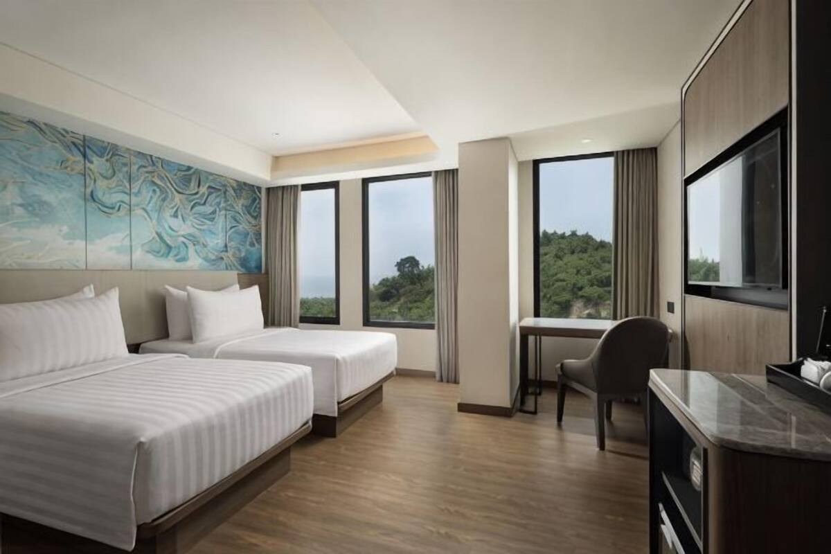 Ein Hotelzimmer mit zwei Betten, einem Schreibtisch, einem Stuhl und großen Fenstern mit Blick auf Grünflächen.