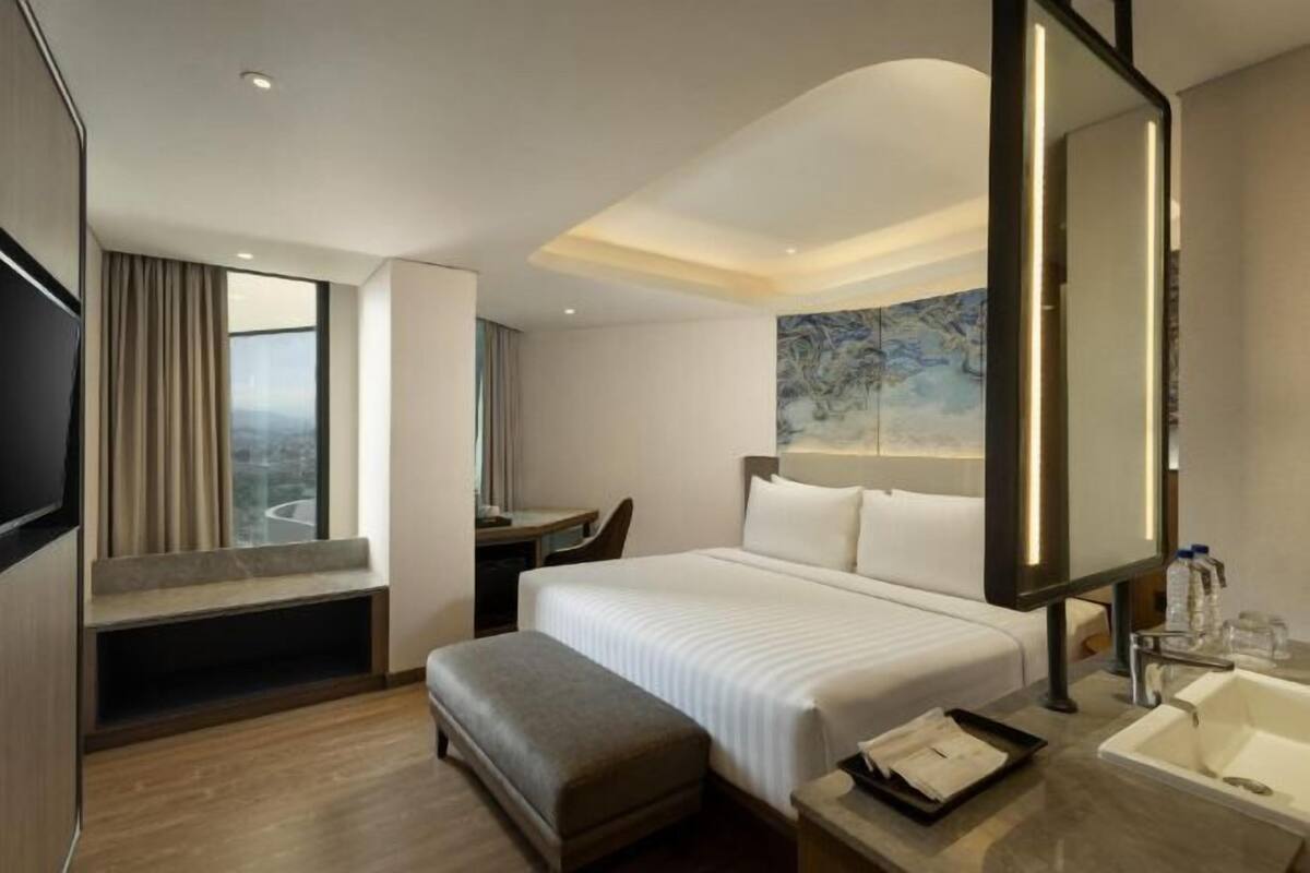 Ein Hotelzimmer mit Bett, Schreibtisch, Stuhl, großem Spiegel, Fenster mit Vorhängen sowie einem Badezimmer mit Waschbecken und Spiegel.