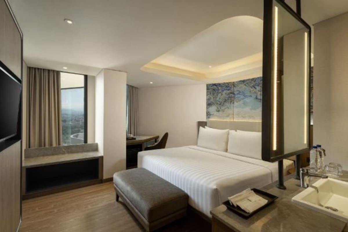 Habitación de hotel con cama, escritorio, silla, un espejo grande, ventana con cortinas y baño con lavamanos y espejo.