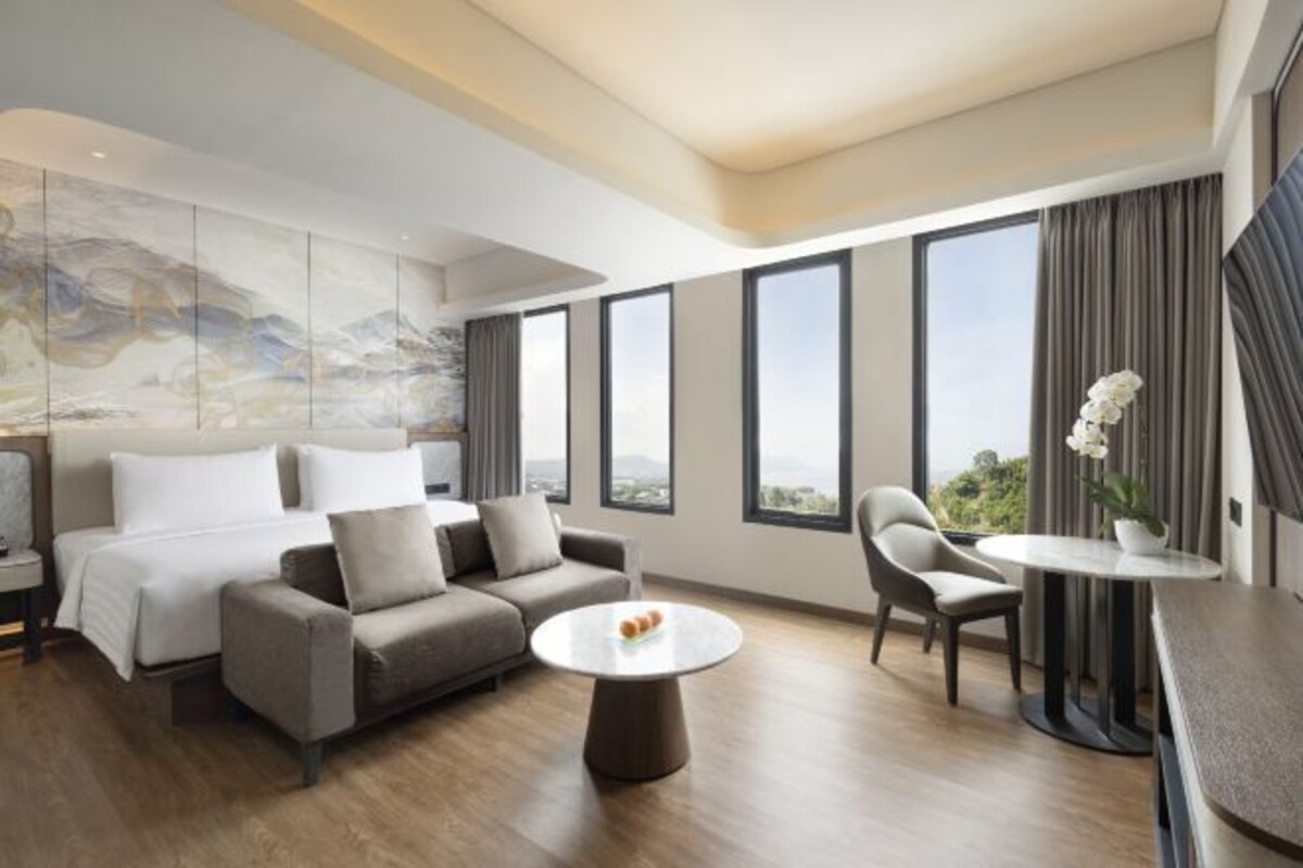 Una habitación de hotel moderna con cama, sofá, sillón, mesa redonda y ventanas amplias que ofrecen vistas a la ciudad.