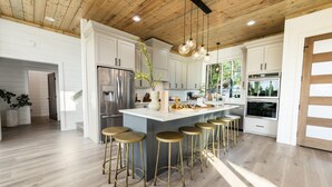 A modern kitchen with white cabinets, a central island with stools, stainless steel appliances, and a wooden ceiling.