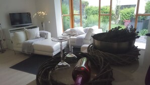 Un salon moderne comprenant un canapé, un téléviseur à écran plat, une table à manger avec des verres à vin, et une vue sur la verdure grâce à de grandes fenêtres.