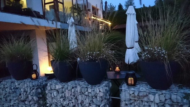 Des plantes en pot, disposées à l’extérieur sur une étagère en pierre, accompagnées d’une table et de chaises, et un parasol blanc en arrière-plan.