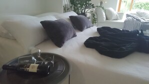 Un lit bien fait, avec du linge de lit blanc et des oreillers foncés, une table de chevet sur laquelle se trouve une bouteille de vin, et une plante en pot à l’arrière-plan.