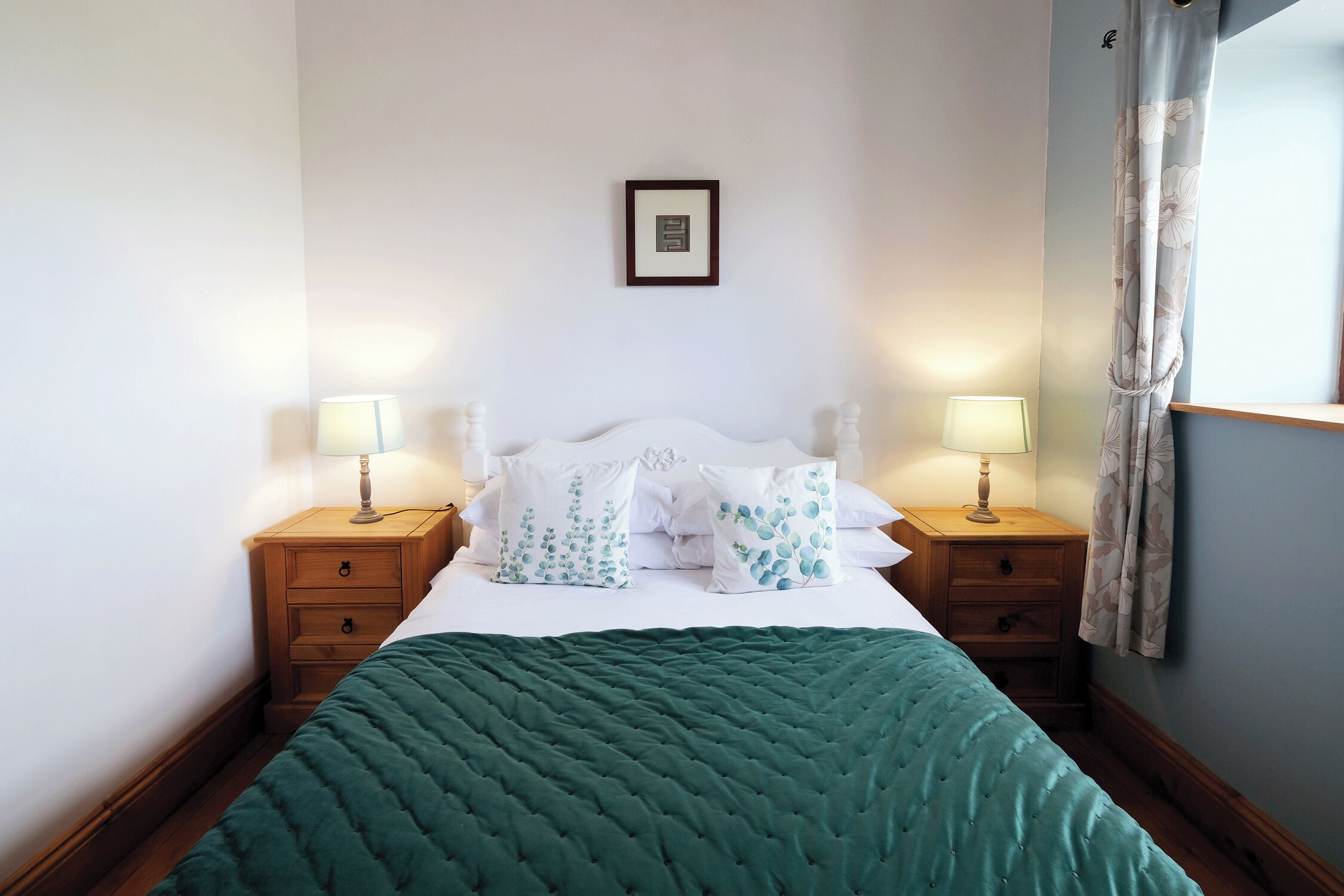 A neatly made bed with a green quilt, two bedside tables with lamps, and a framed picture on the wall.