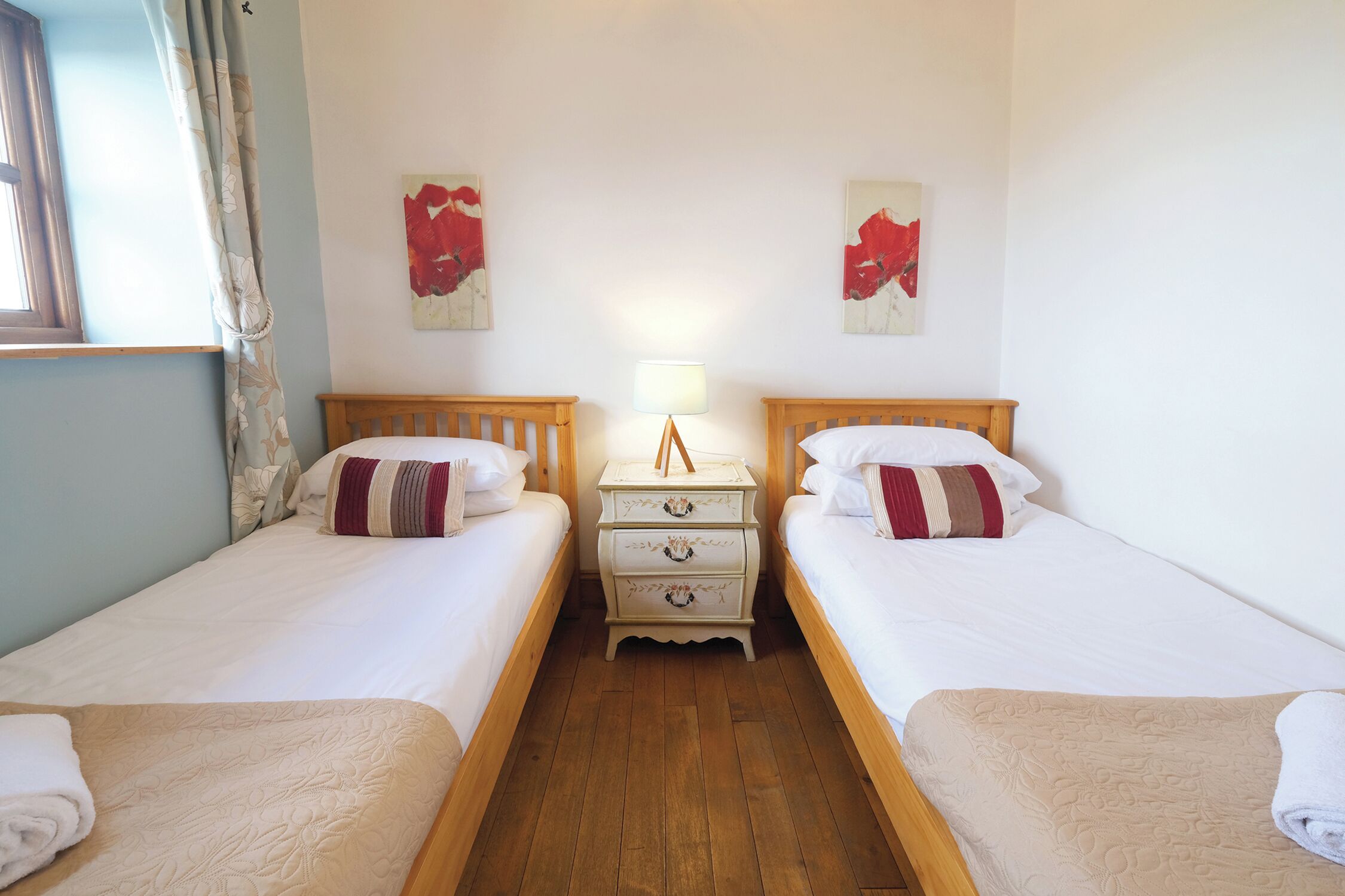 A room with two single beds, a nightstand with a lamp, and artwork on the walls.