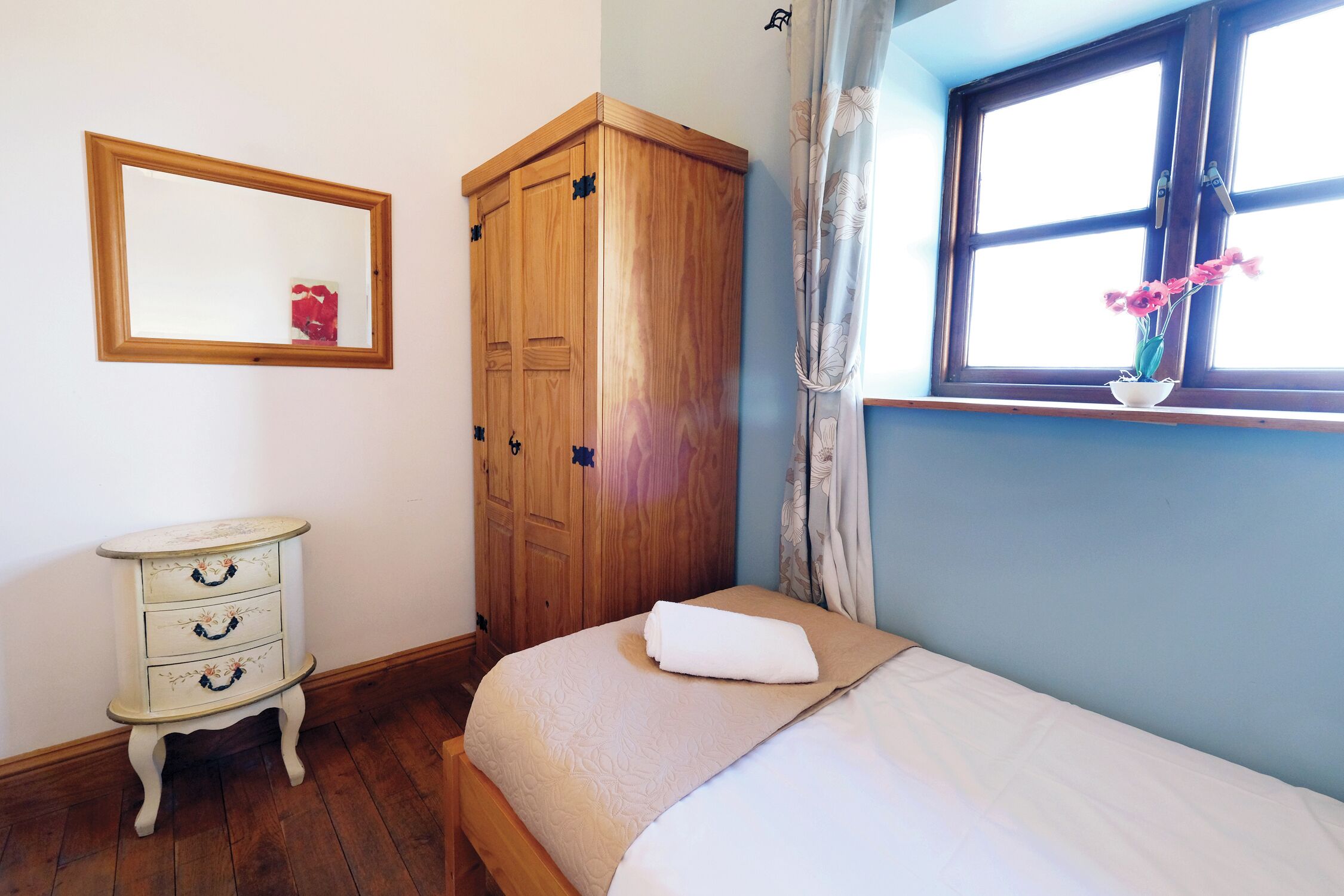 Une chambre à coucher avec un lit, une armoire en bois, une petite commode blanche, un miroir et une fenêtre avec un rideau.
