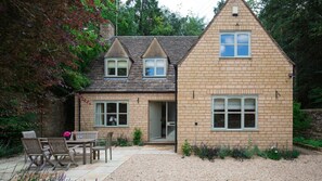 A two-story house with a brick exterior, a small front porch with wooden chairs, and a gravel driveway. - Woodland Cottage - Temple Guiting, Dog Friendly (Temple Guiting)