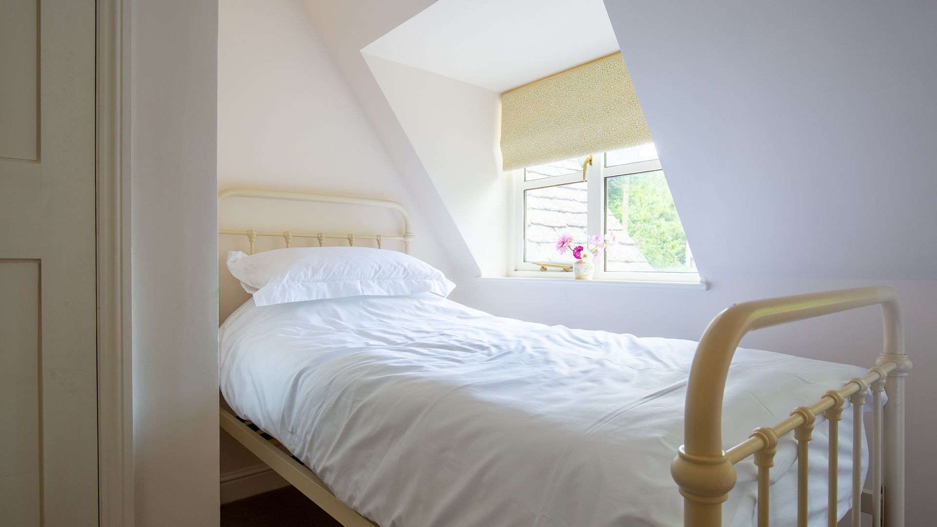 Ein Einzelbett in einem Zimmer mit schrägen Wänden und einem Fenster mit gemustertem Rollo.