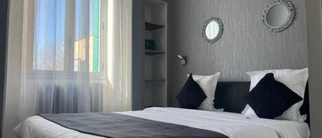 Un dormitorio con cama, ventana con cortinas traslúcidas, armario y dos espejos adosados a la pared.