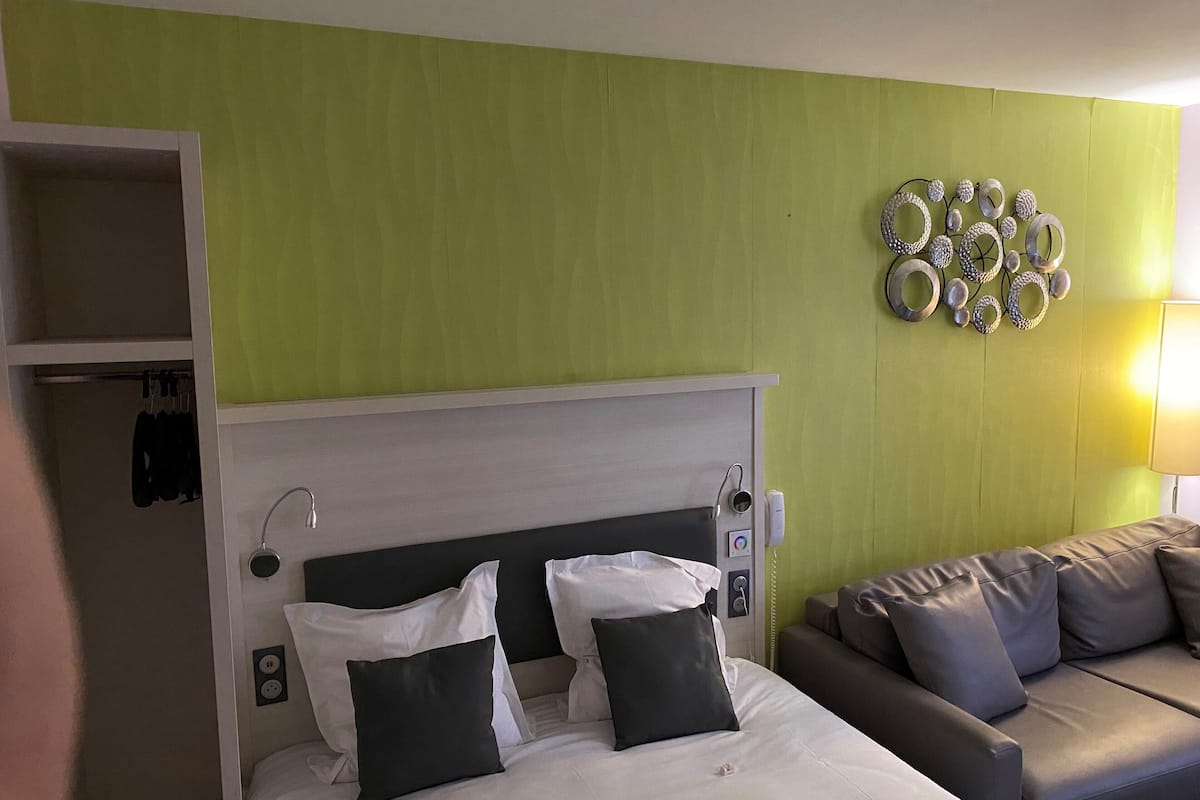 A hotel room with a bed, a grey sofa, a green wall, and a decorative metal piece.