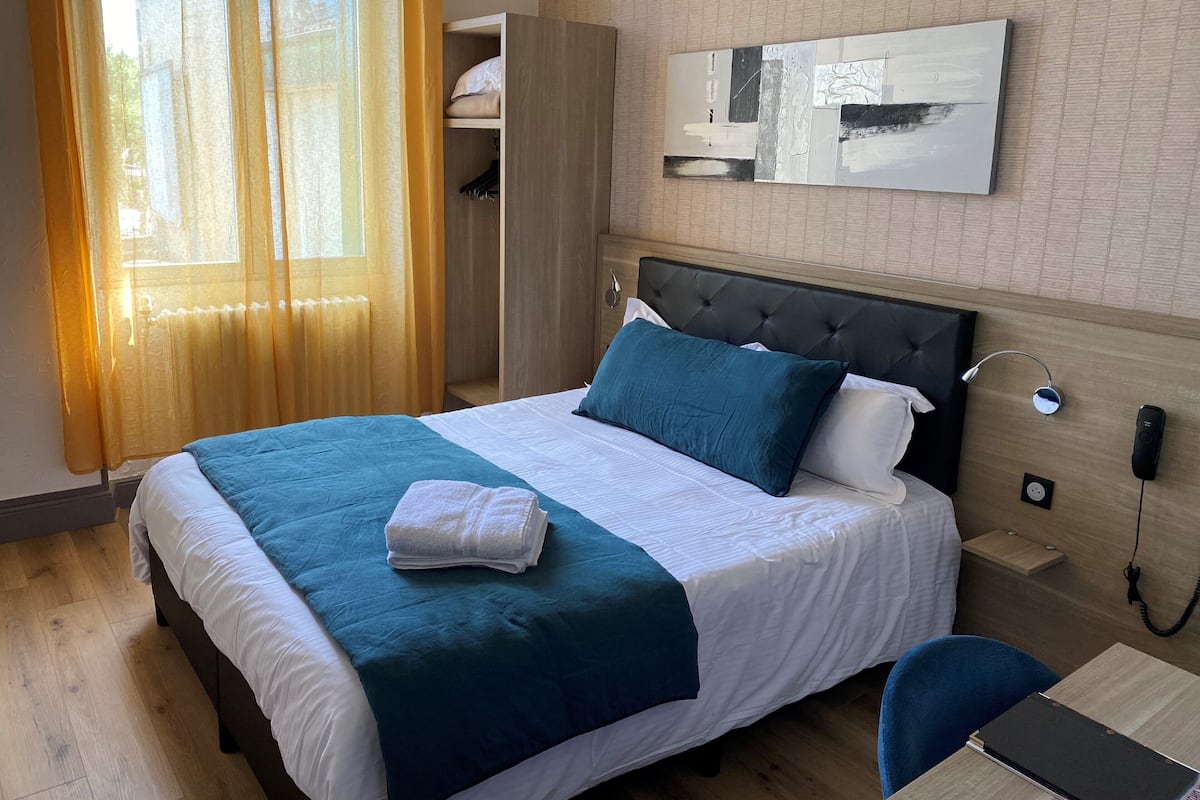 A hotel room with a bed, a bedside table, a desk, a chair, a wardrobe, and a window with curtains.