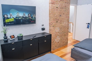 Ein modernes Zimmer mit einem Fernseher, der die Serie 'Brooklyn Nine-Nine' zeigt, einer schwarzen Kommode mit einer Pflanze und einer Ziegelwand.