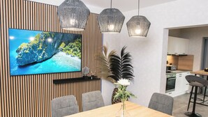 A modern dining area with a wooden table, grey chairs, a TV mounted on a wooden panel wall, and pendant lights.