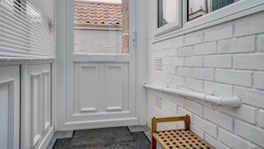 A narrow hallway with a white door, a wooden bench, and a white brick wall with a window and a pipe.