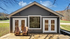 A small, single-story building with a gray exterior and white trim. It features two large windows and two doors, each with a small light above. There is a wooden deck with two chairs and a table in front. - Getaway Cottage, Pickleball Court, Creek, WIFI, Mountain Views, Starting at $75 (Cosby)