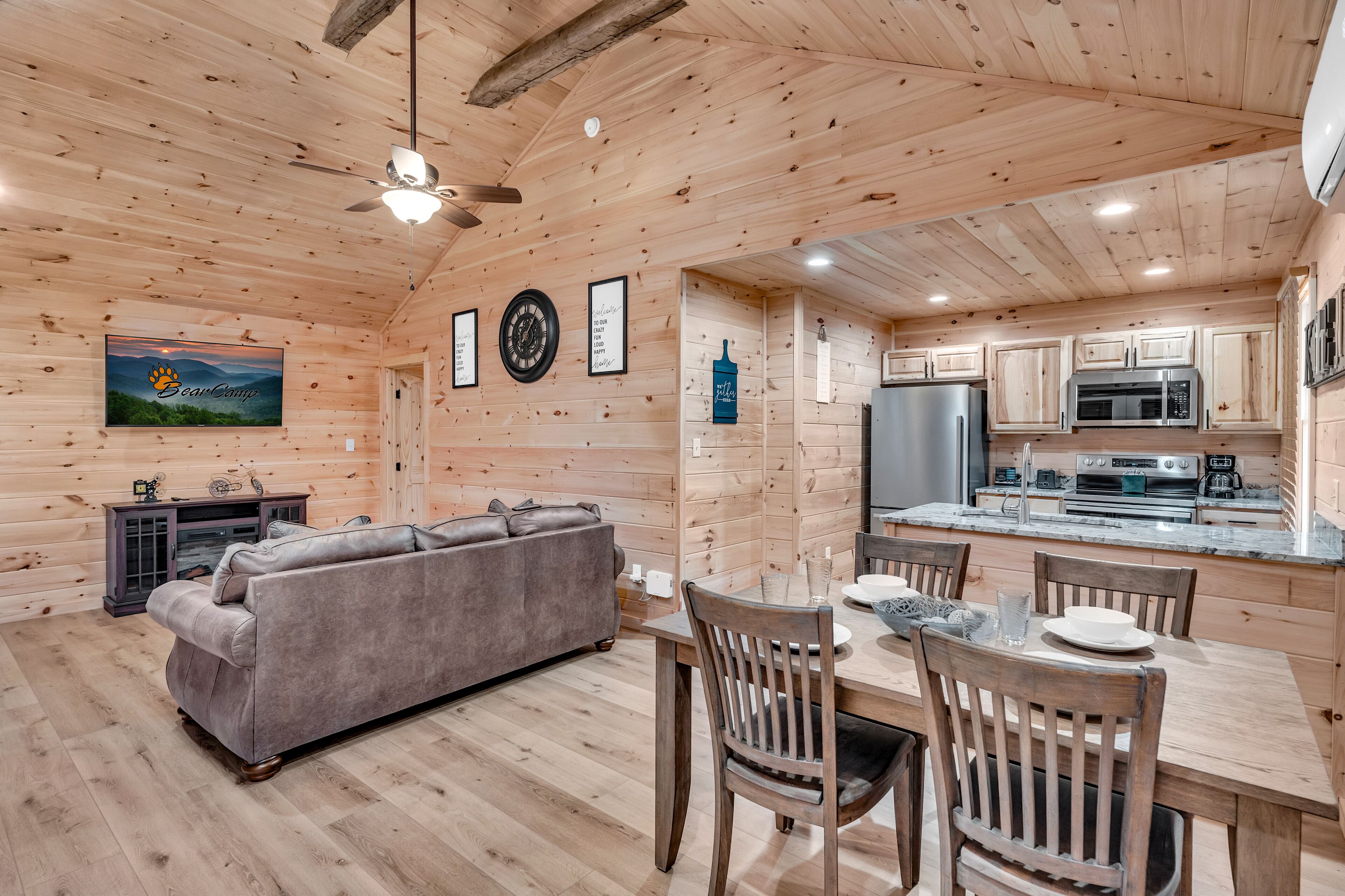 A wooden cabin interior with a sofa, dining table, chairs, a ceiling fan, a TV, framed pictures, and a kitchen area with appliances.