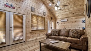 L’intérieur d’une cabane en rondins, avec un plancher en bois, un canapé en cuir marron, une table basse en bois et un ventilateur de plafond.