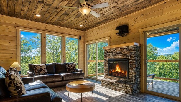 L’intérieur d’une cabane en rondins, avec une cheminée en pierre, un canapé en cuir et de grandes fenêtres.