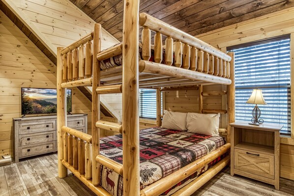 Un lit superposé avec une structure en bois et une couvre-lit matelassé, une table de chevet avec une lampe et une commode ornée d’un tableau représentant un paysage.
