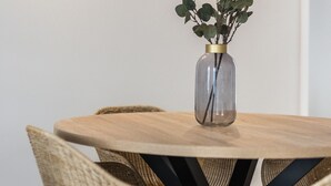 A round wooden table with wicker chairs, a glass vase with green foliage, and wall-mounted lights. - Suits Boutique Arco - Mezanino apartment by HD (Faro)