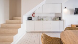 A modern kitchen with white cabinetry, a microwave, and a coffee maker. There is a staircase leading to another level and a dining area with a wooden table and wicker chairs. - Suits Boutique Arco - Mezanino apartment by HD (Faro)