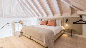 Una camera da letto con soffitto a falde, un letto con copripiumino color beige, due cuscini, un piccolo tavolino rotondo e un punto luce fissato al muro.