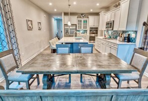 A dining area with a large wooden table and blue chairs, a kitchen with white cabinets and blue accents, and artwork on the walls.