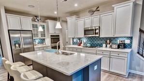 A modern kitchen with white cabinetry, a large island with bar stools, stainless steel appliances, and a blue backsplash.