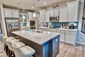 A modern kitchen with white cabinetry, a large island with bar stools, stainless steel appliances, and a blue backsplash. - Bayside * 4BR Luxury Twinhouse (Selbyville)