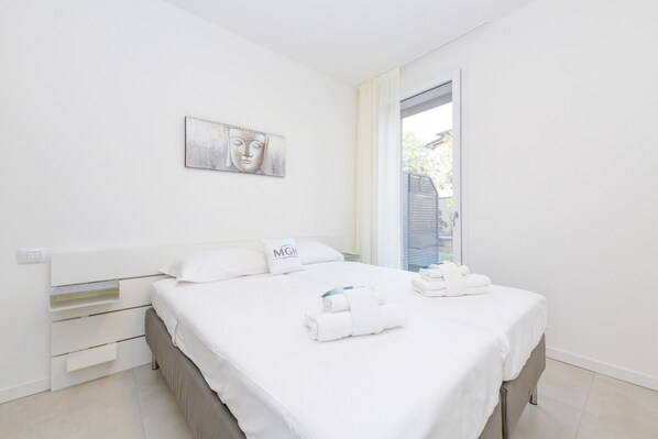 A modern hotel room with a large bed, a bedside table, a wall-mounted artwork, and a view through a window.