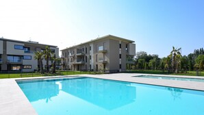 A swimming pool with a clear blue surface, surrounded by a paved area and a multi-story building with balconies.