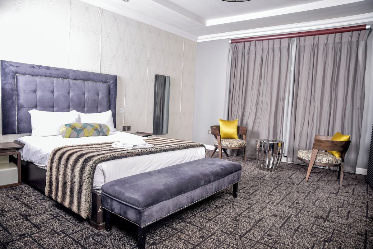 A hotel room with a large bed, a bench, two chairs, a small table, and a large window with curtains.