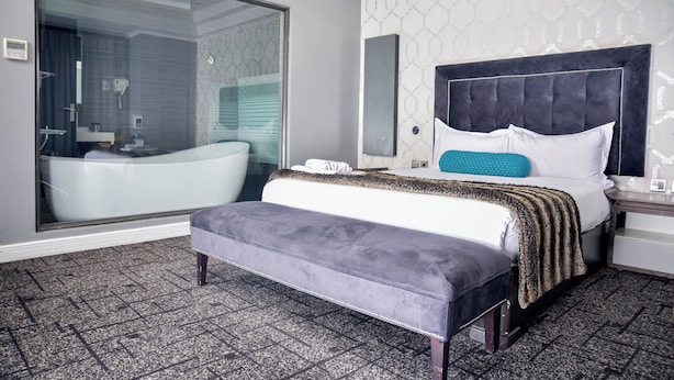 Een moderne hotelkamer met een ruime, glazen badkamer, een gestuikt hoofdeinde, een groot bed met een bedsprei met een patroon, en een bankje.