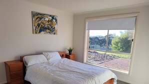 Une chambre à coucher avec un lit, des tables de chevet en bois, une plante en pot et une grande fenêtre donnant sur un jardin et des bâtiments.